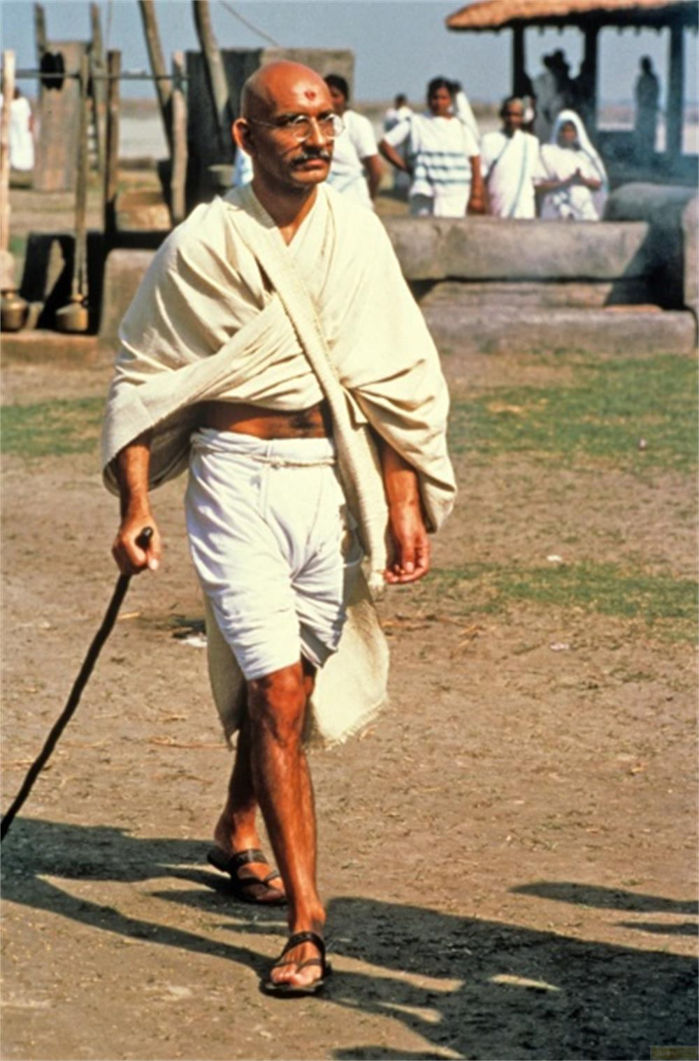 Un homme chauve avec des lunettes marche avec une canne. Il porte un tissu blanc et un short.