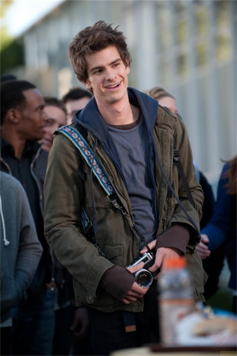 Un jeune homme en veste verte et sweat à capuche bleu sourit, tenant un appareil photo avec une sangle à motifs.