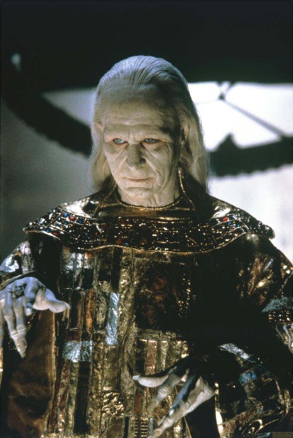 Un vieil homme aux longs cheveux blancs et yeux bleus porte une robe dorée ornée.