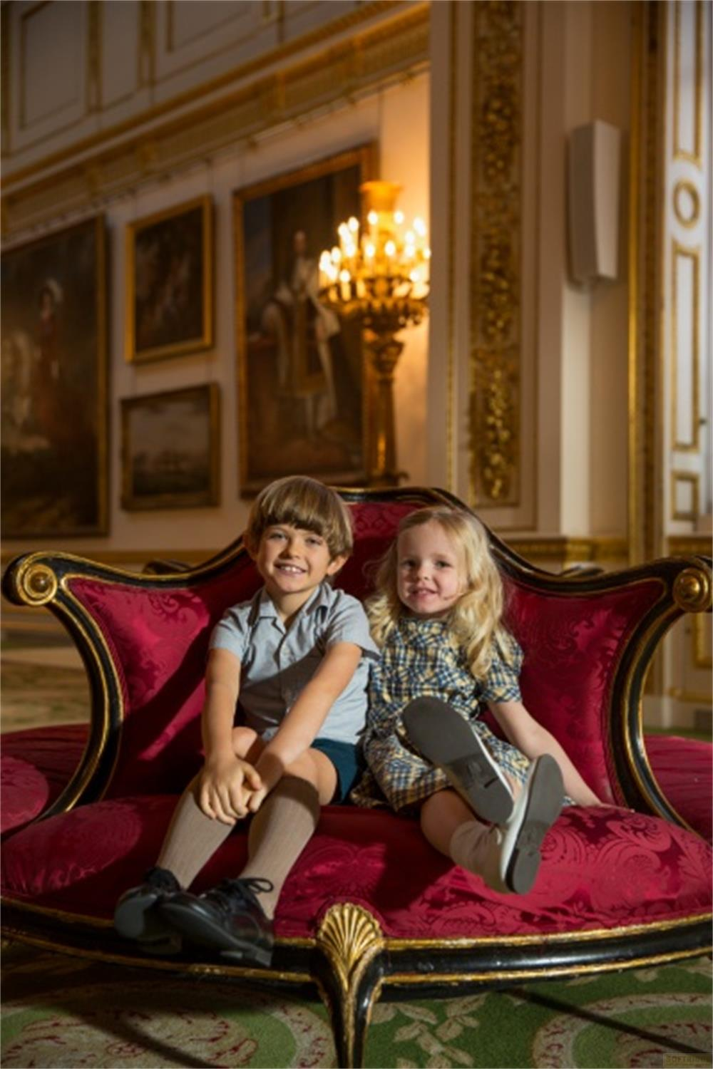 Due bambini siedono su un divano di velluto rosso in una stanza opulenta con dipinti.