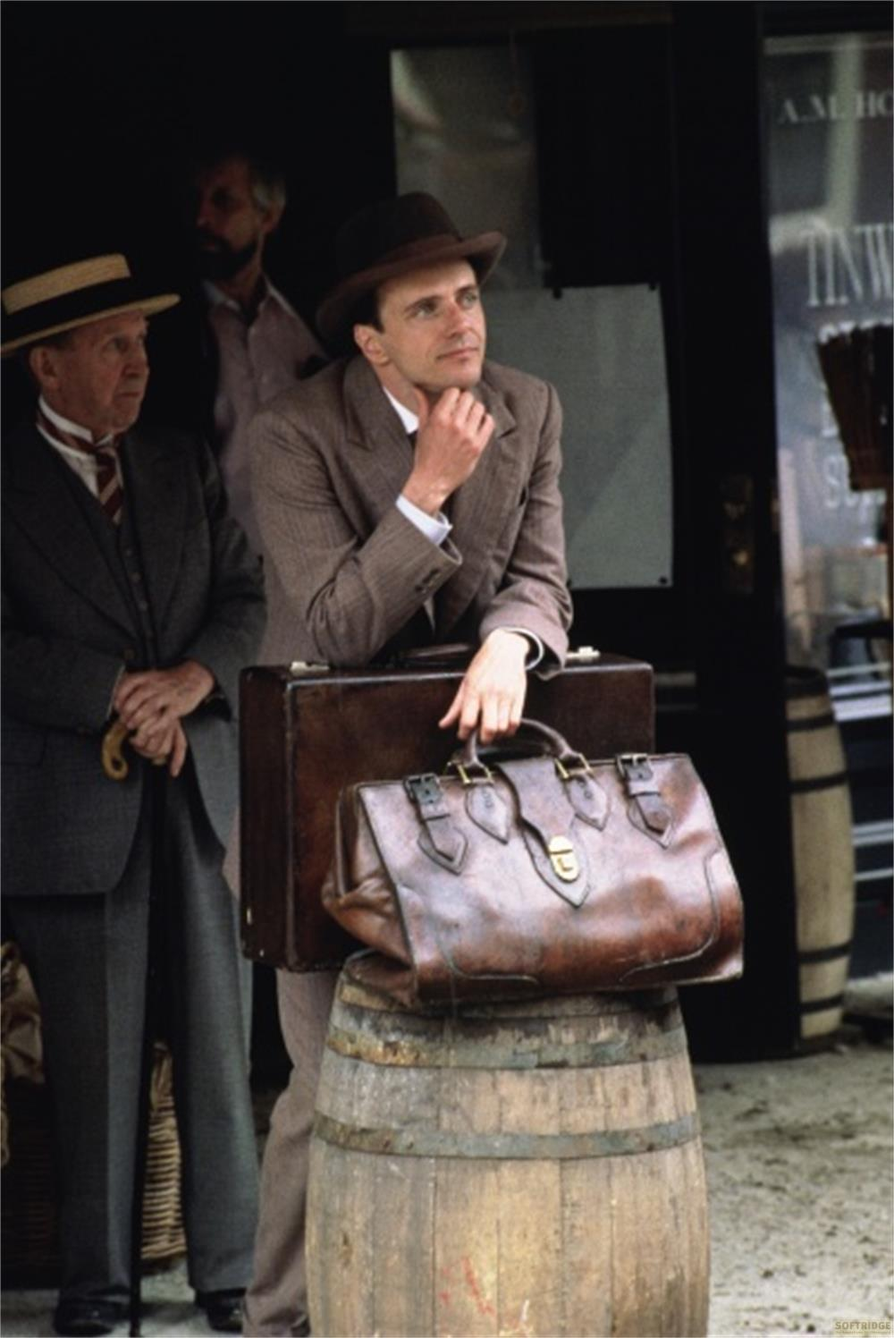 Due uomini con cappelli e abiti stanno con bagagli vintage su un barile di legno.