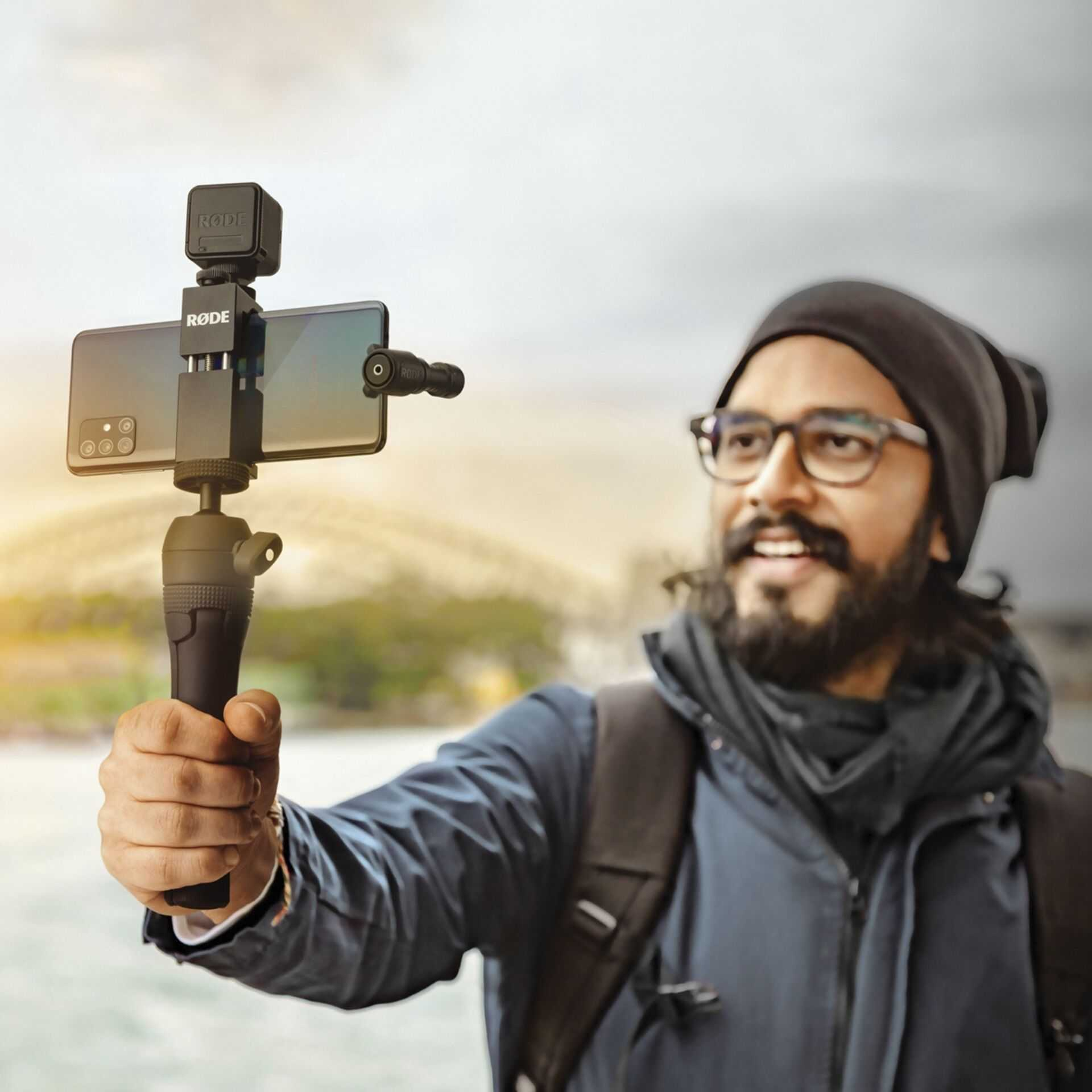 Uomo con occhiali e barba tiene smartphone con microfono e treppiede.