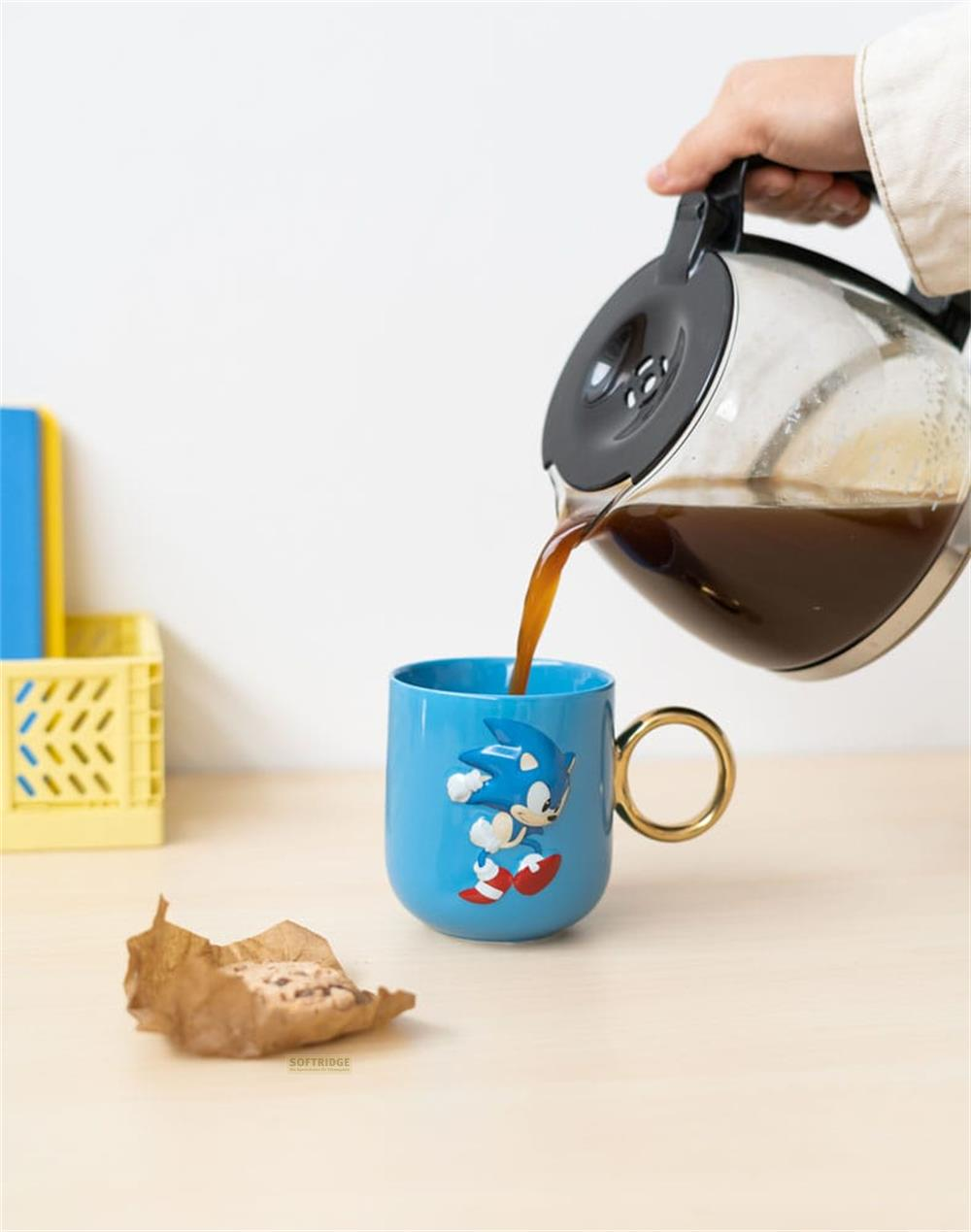 Kaffee wird in eine blaue Sonic-Tasse gegossen, daneben liegt ein zerknülltes Papier.