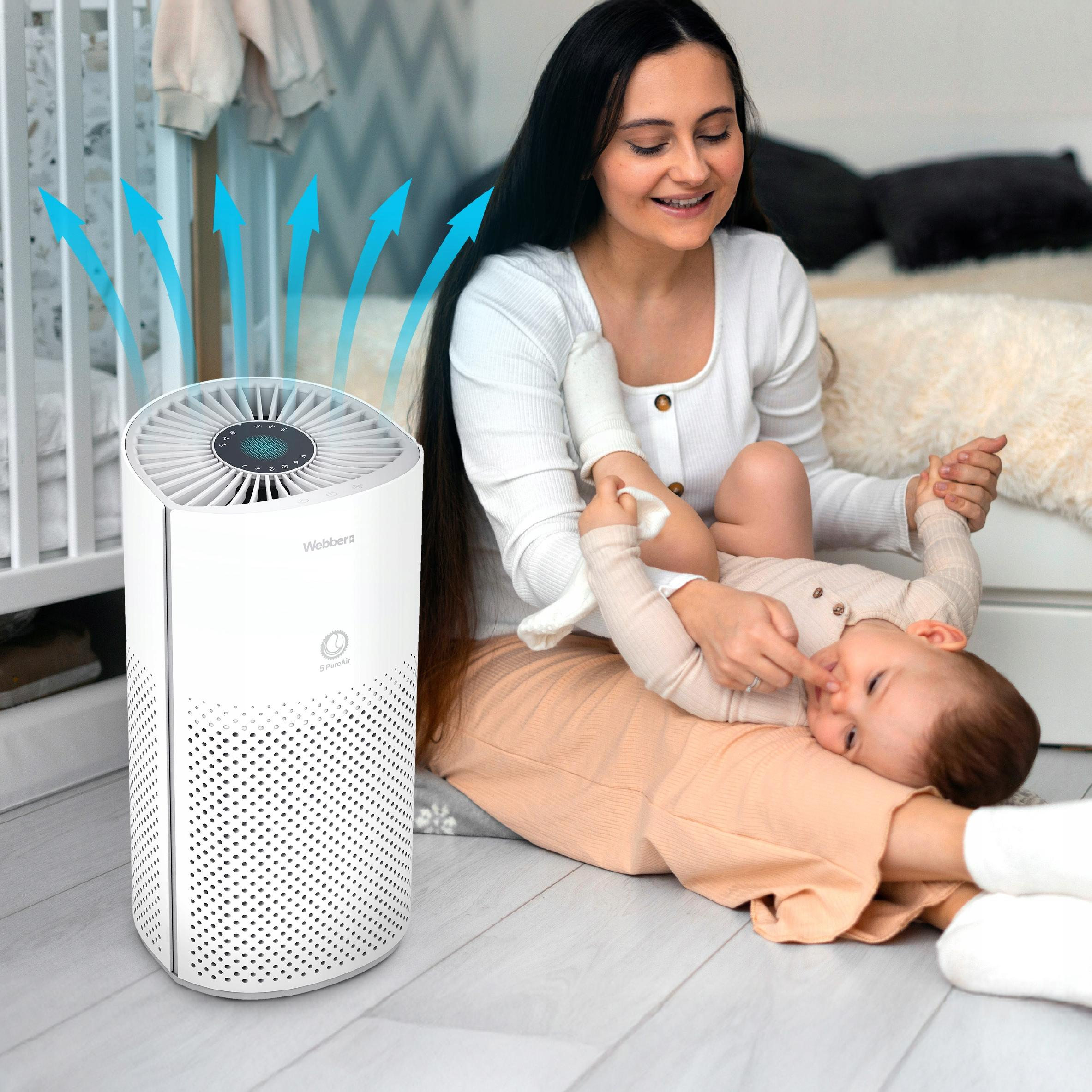 Biały oczyszczacz powietrza obok łóżeczka; kobieta trzyma dziecko.