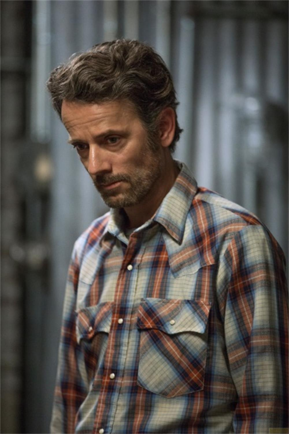 Un uomo con la barba indossa una camicia a quadri. Guarda in basso.