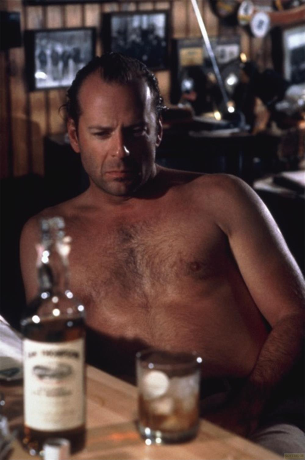 Un homme chauve est assis torse nu devant une bouteille et un verre de whisky.
