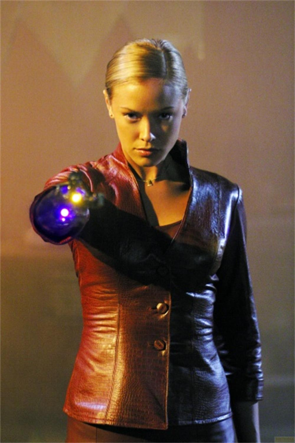 Une femme blonde en veste de cuir bicolore vise avec un orbe lumineux violet et jaune de sa main gauche, sérieuse.