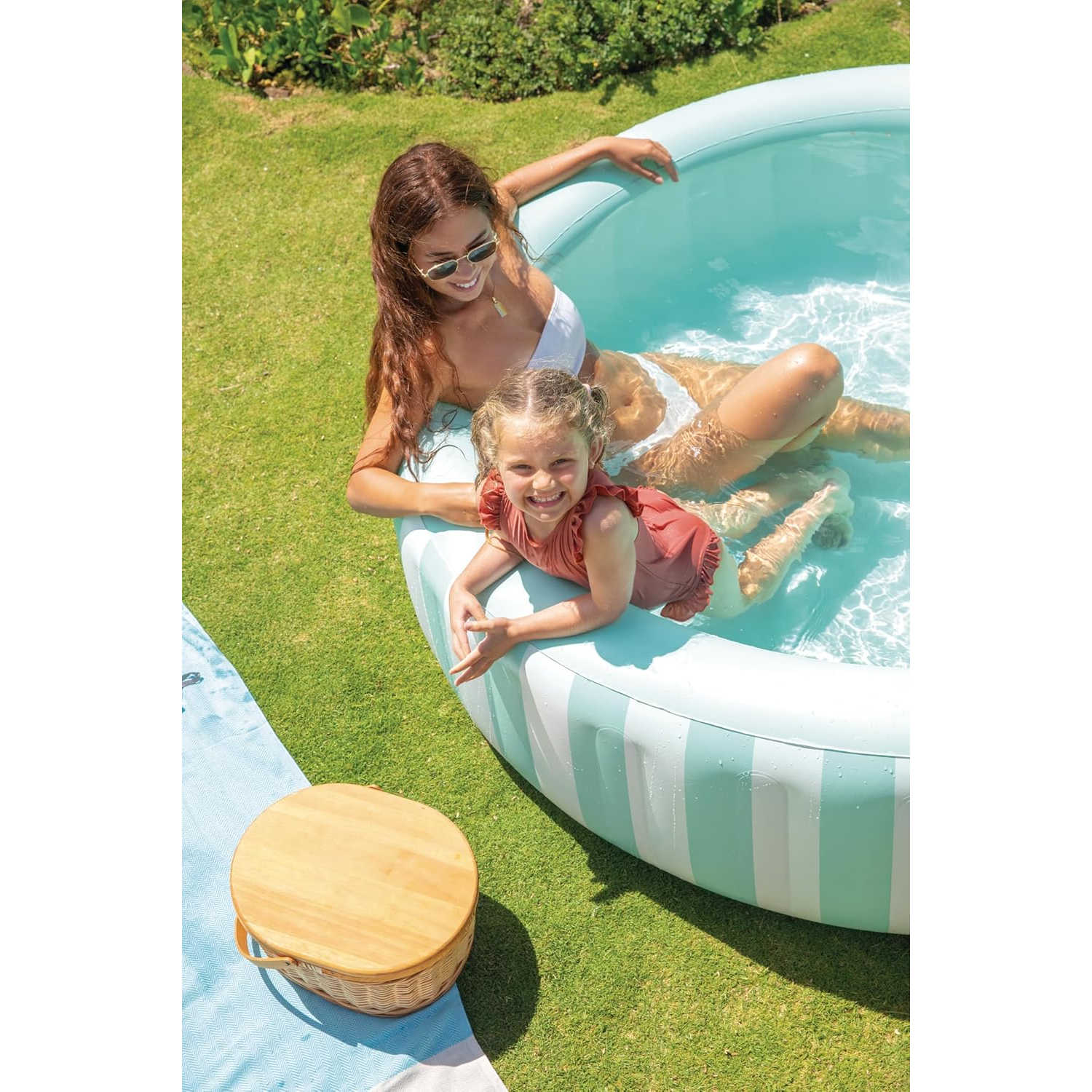 Eine Frau und ein Kind planschen in einem hellblauen Pool auf einer grünen Wiese.