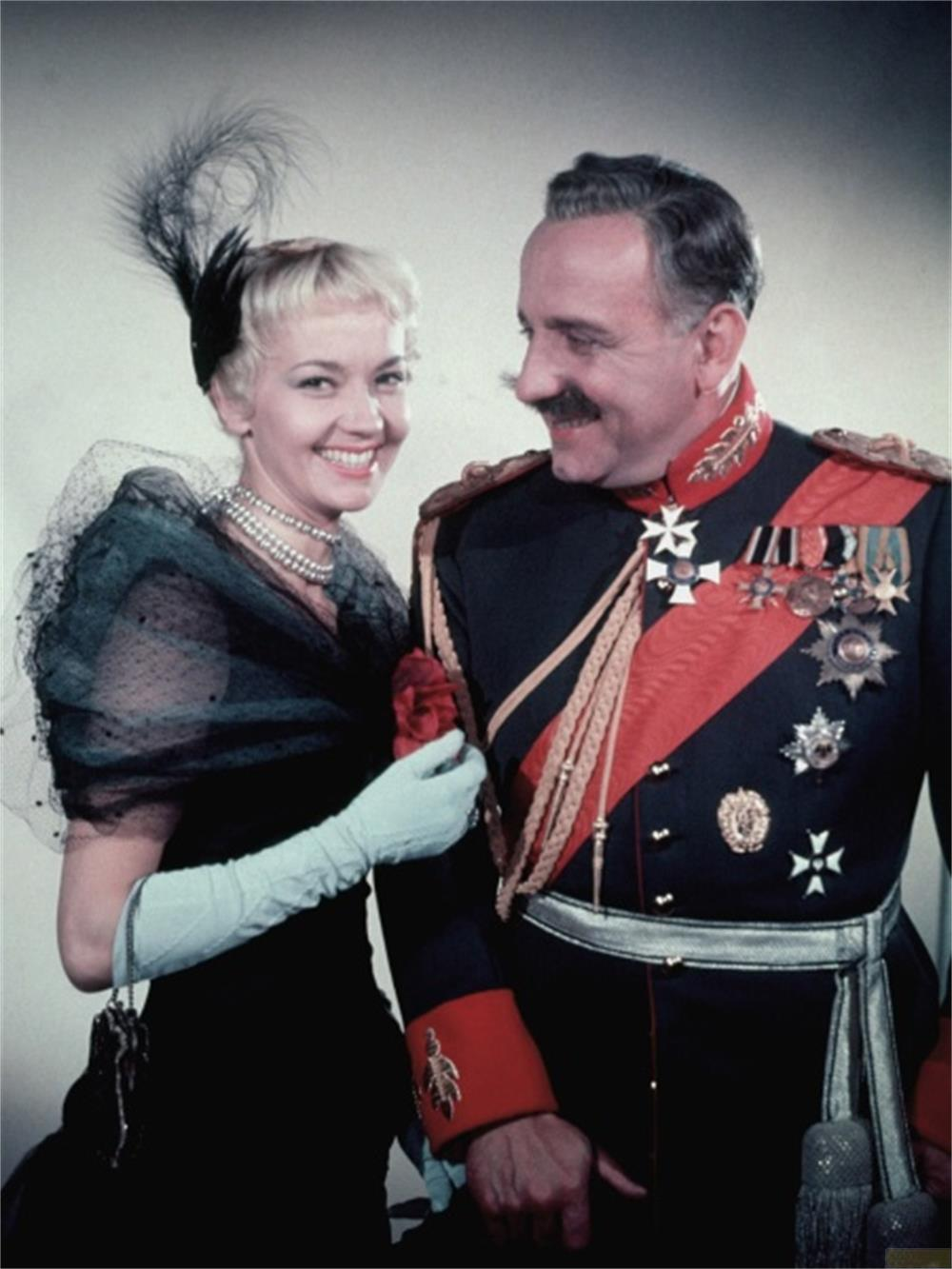 Ein Mann in Uniform und eine Frau in schwarzem Kleid mit Hut und Handschuhen.