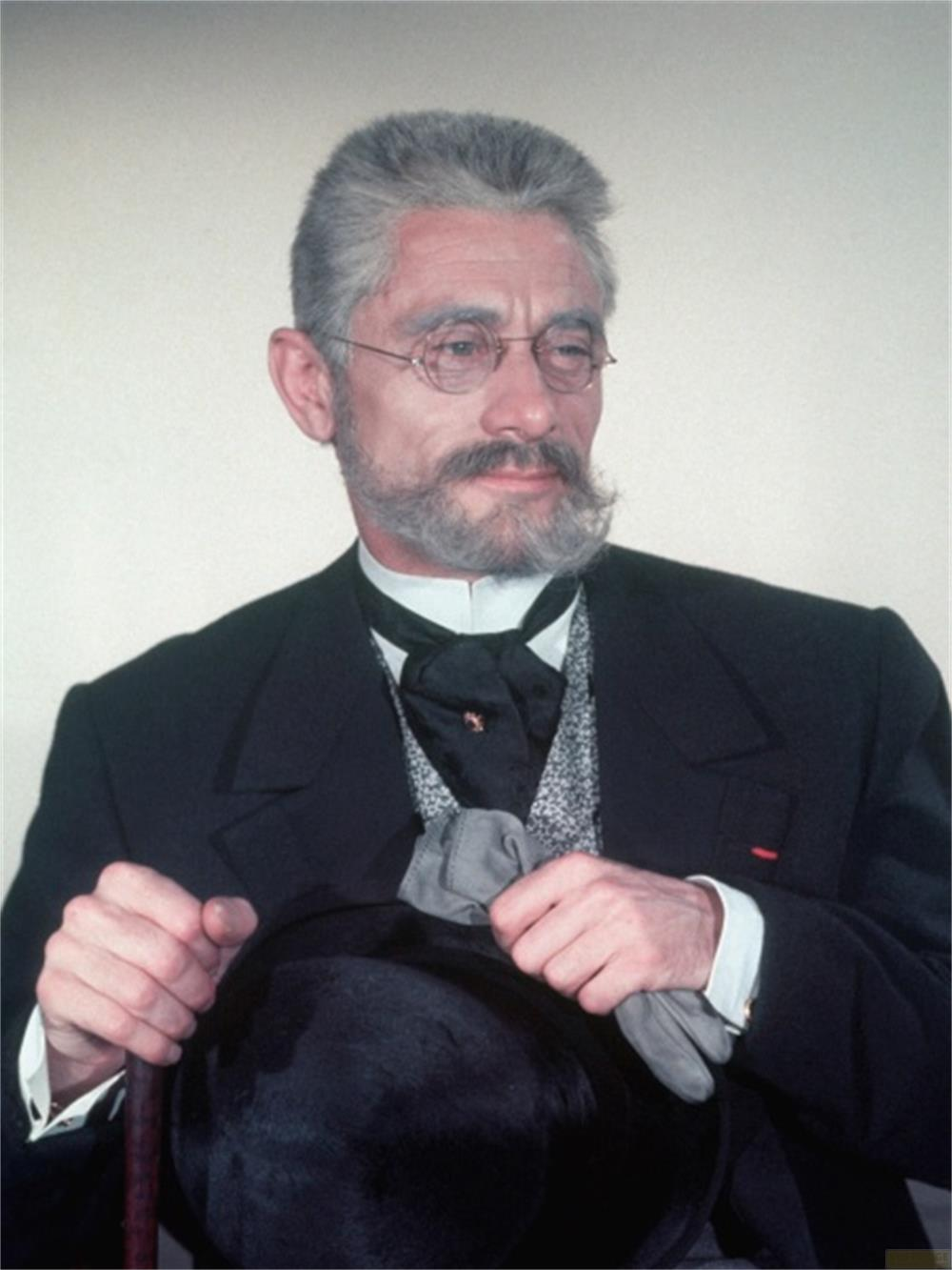 Ein Mann mit Brille, Schnurrbart und grauem Haar hält einen Hut und einen Stock.