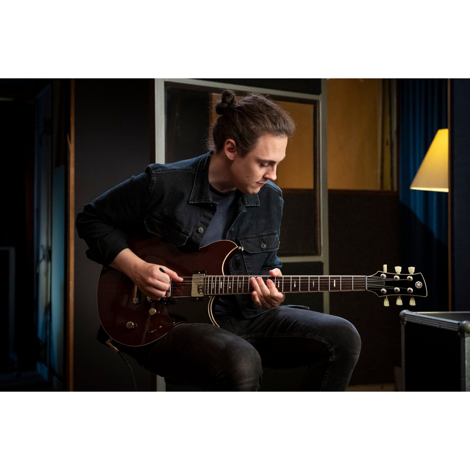 A man with a bun plays a brown electric guitar in a dark room.