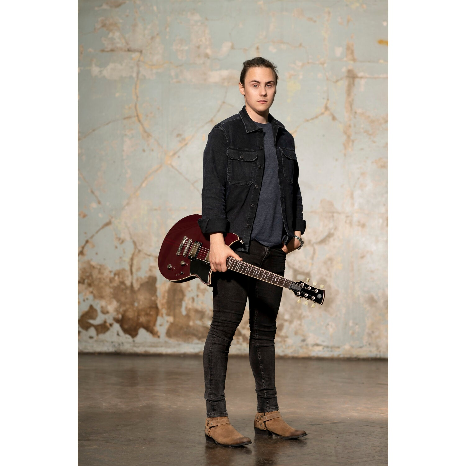 A man in a dark shirt holding a guitar stands against a textured wall.