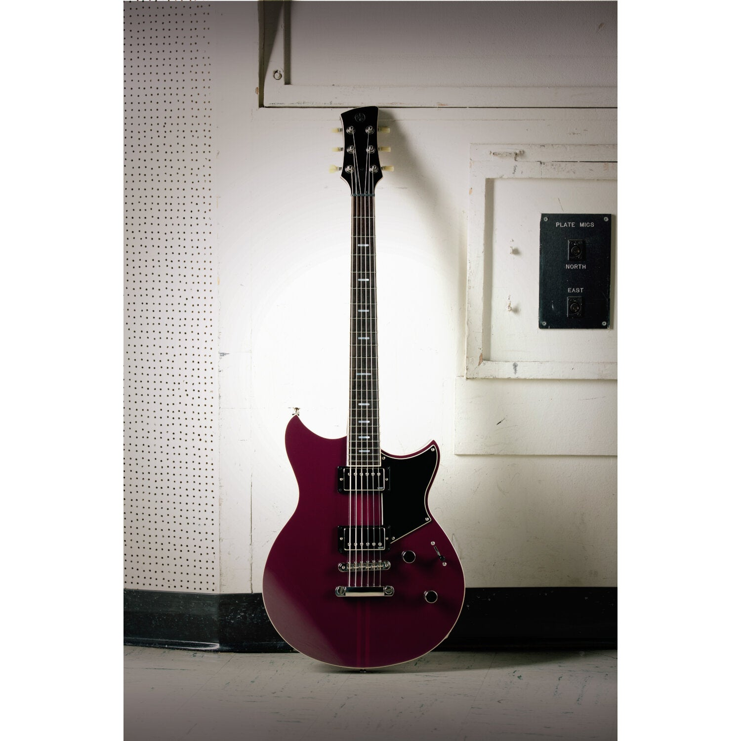 A burgundy electric guitar stands upright against a wall with pegboard and switches.