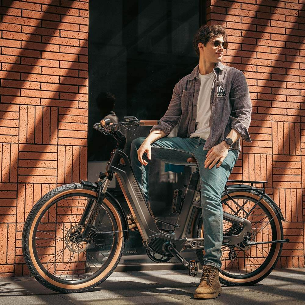 Mężczyzna siedzi na rowerze elektrycznym przed ceglanym murem. Nosi dżinsy i koszulę.