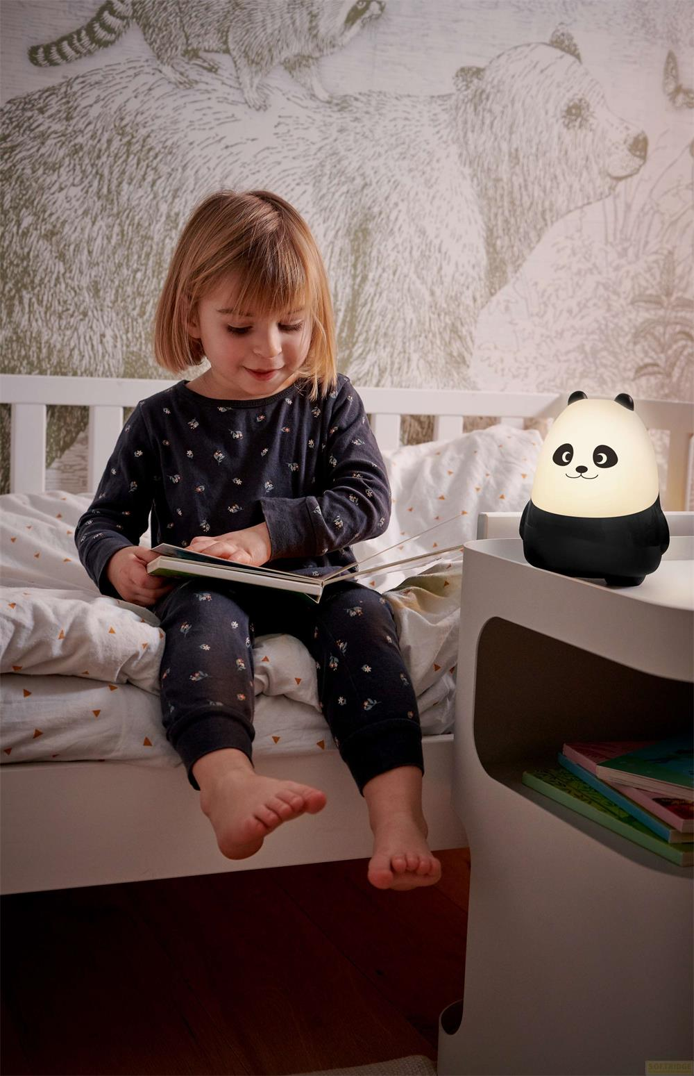 Une fille lit un livre au lit, avec une lampe panda lumineuse à côté.