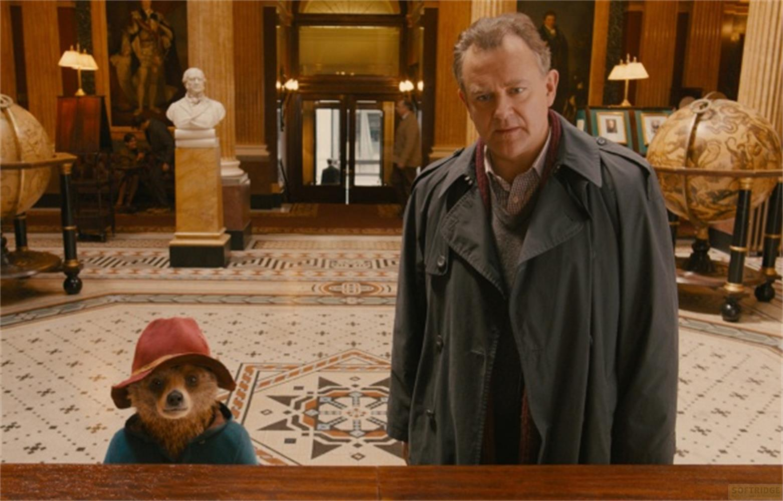 Un orso con cappello rosso e un uomo in cappotto in un atrio.