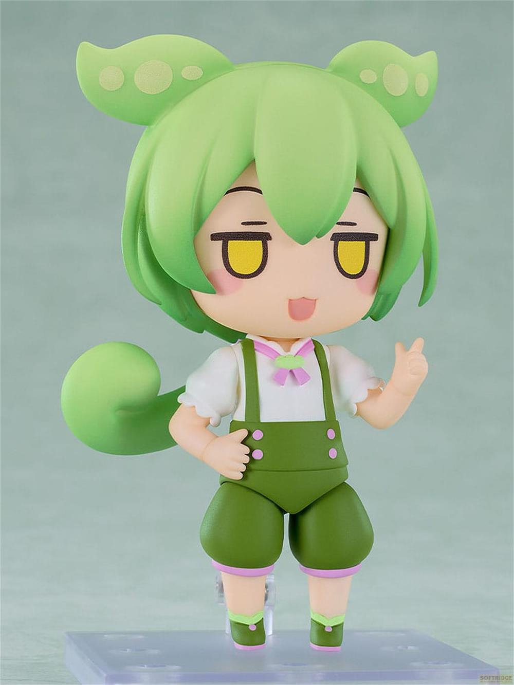 Grüne Figur mit Latzhose und Schleife. Hält die Hand erhoben.