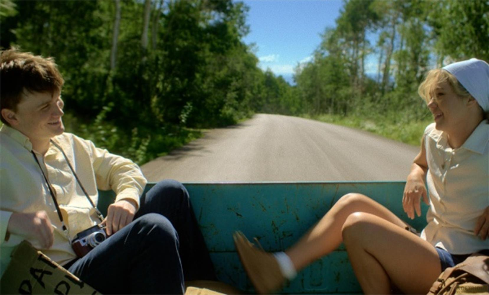Una coppia viaggia sul retro di un pick-up lungo una strada, circondata da alberi.