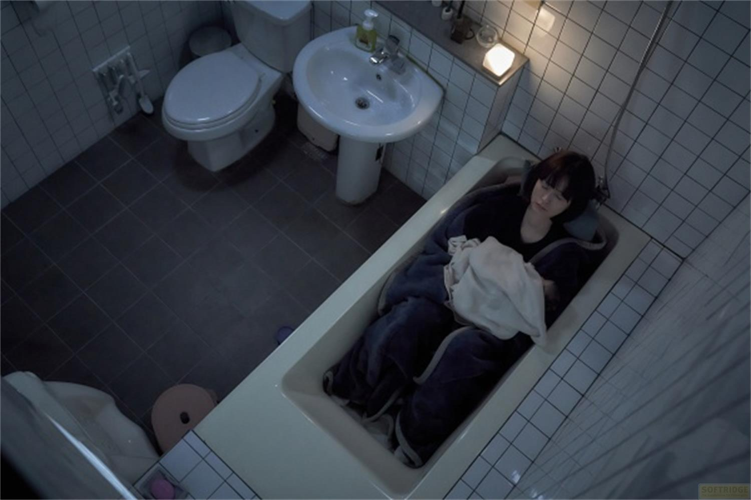 Una donna giace in una vasca da bagno coperta da coperte. Toilette e lavandino sullo sfondo.