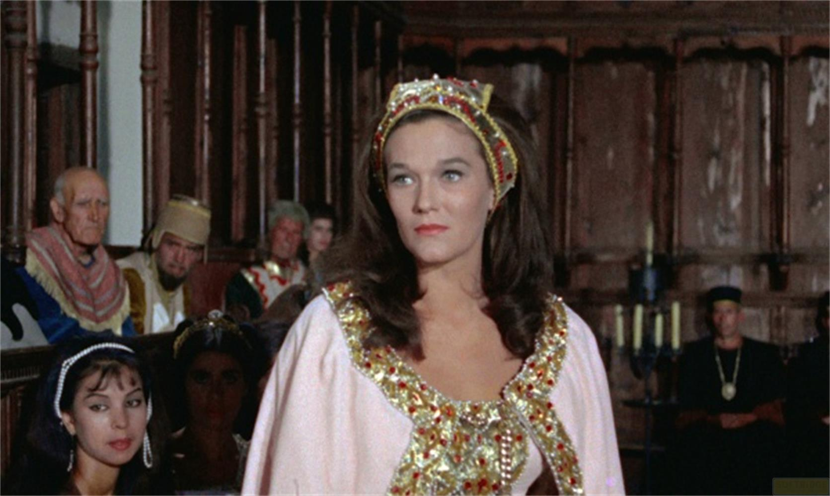 Una donna con corona indossa un abito rosa con finiture dorate. Persone sedute sullo sfondo.