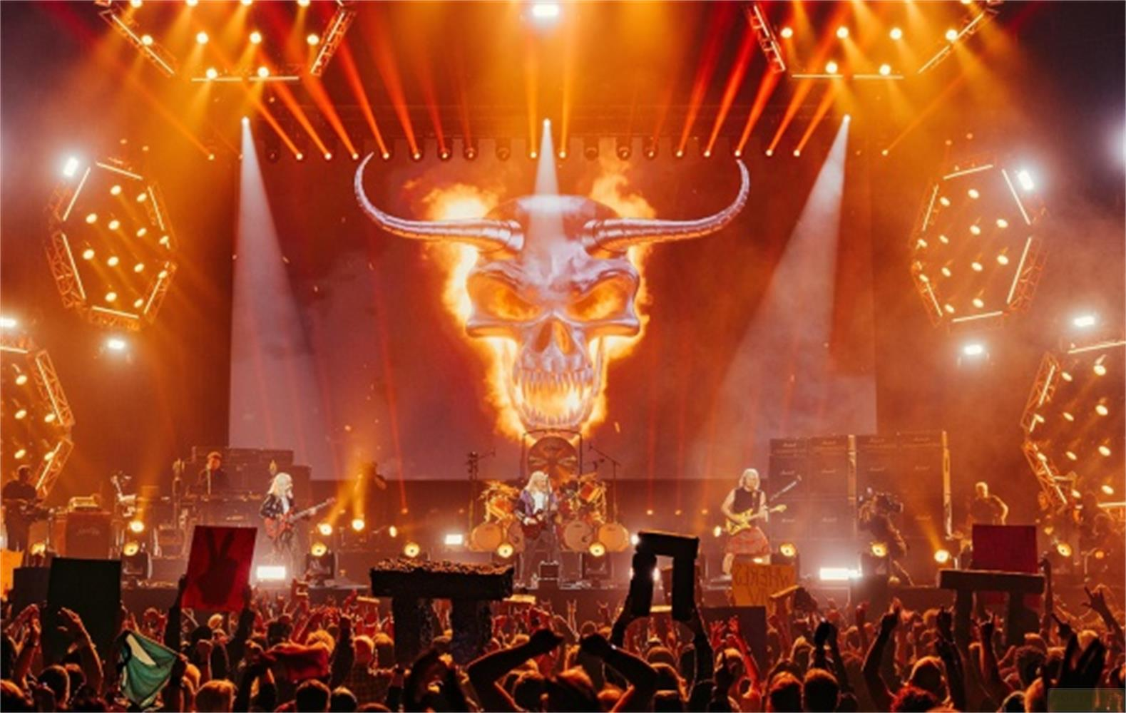 Bühnenkonzert mit Totenkopf-Logo und Feuer, Band spielt vor jubelndem Publikum.