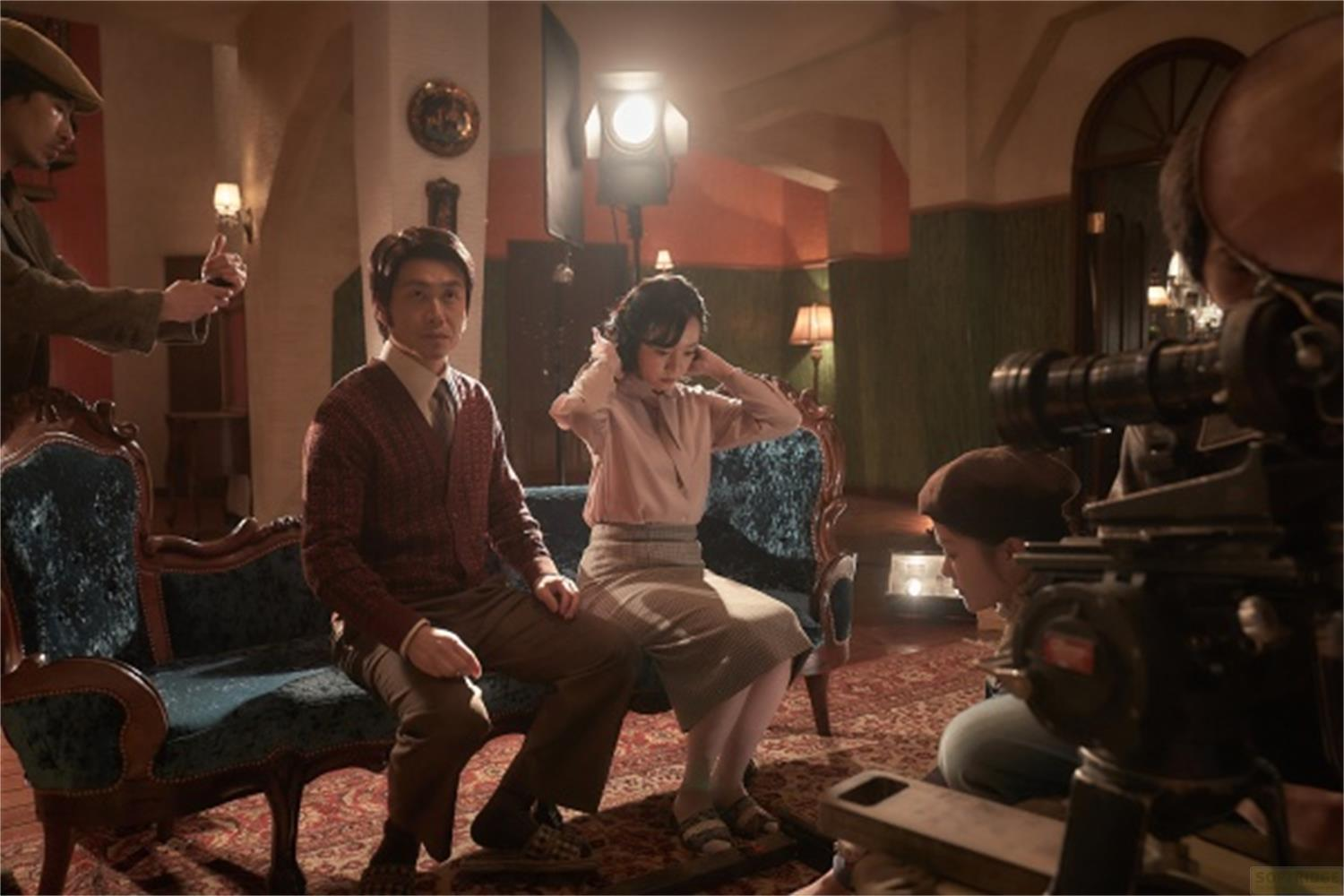 Un uomo e una donna siedono su un divano mentre una troupe cinematografica riprende.