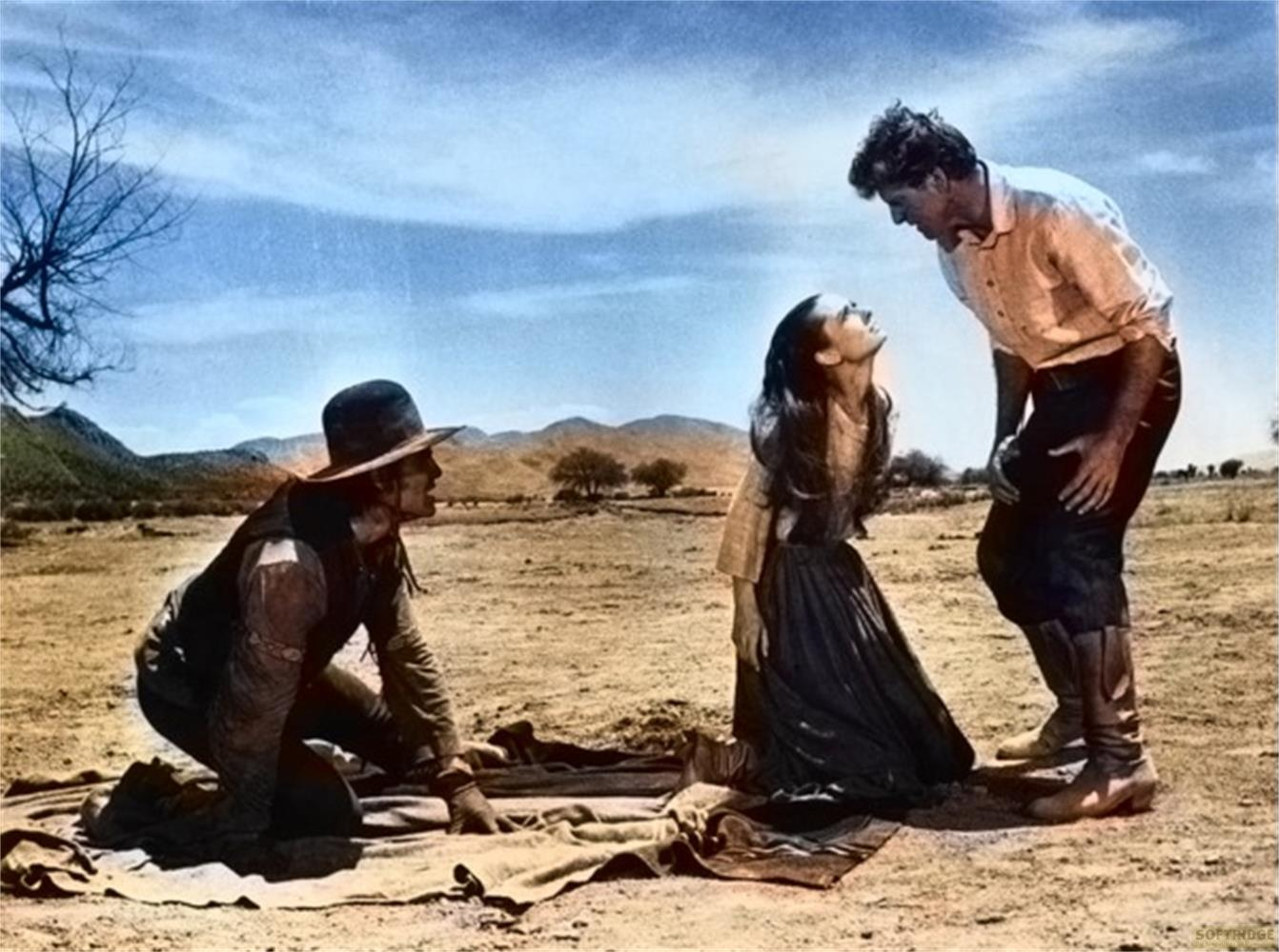 Un uomo in ginocchio davanti a una donna che lo guarda. Un altro uomo vicino.