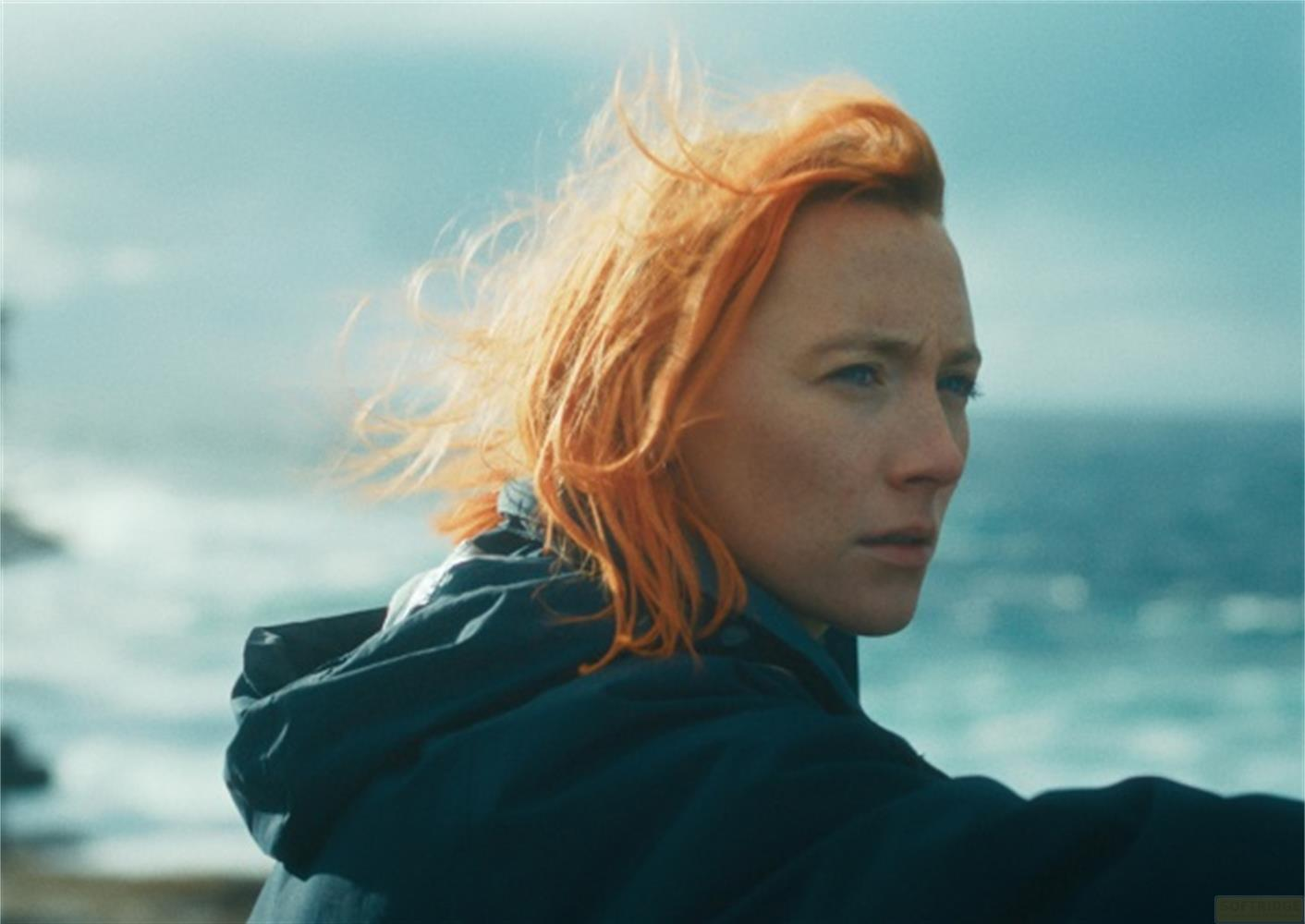 Frau mit roten Haaren blickt aufs Meer, trägt dunkle Jacke.
