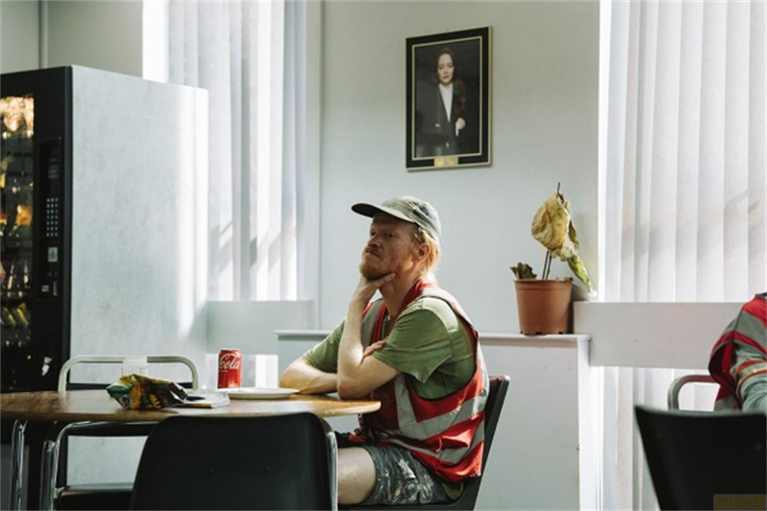 Uomo barbuto seduto a tavolo, beve cola, indossa gilet. Ritratto sul muro.