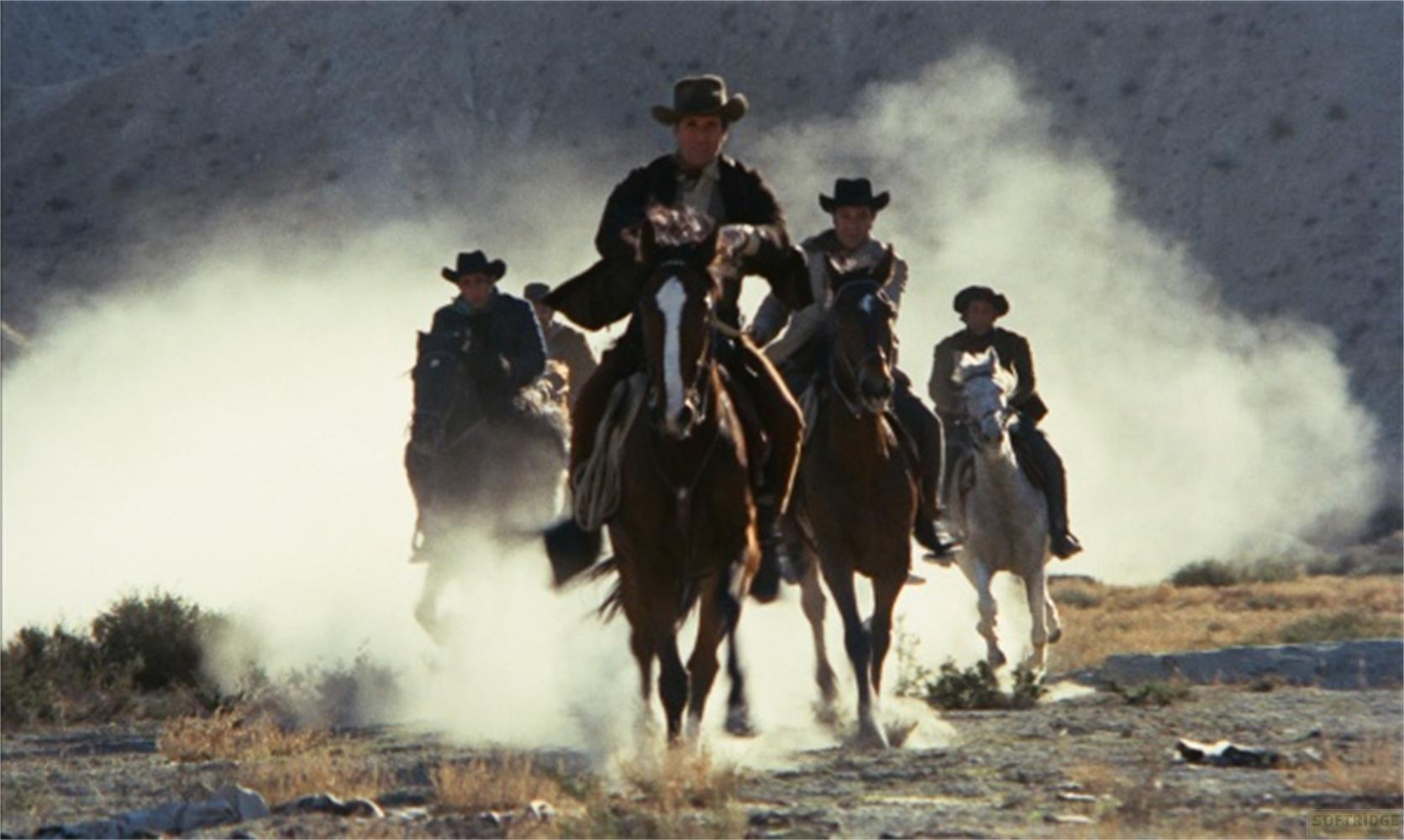 Quattro cowboy a cavallo cavalcano in un deserto polveroso. Montagne sullo sfondo.