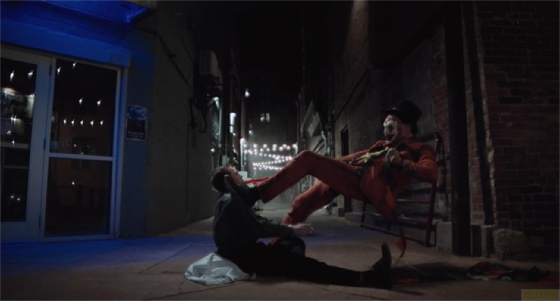 Un uomo in completo rosso, cappello a cilindro e trucco da clown prende a calci un altro uomo in un vicolo.