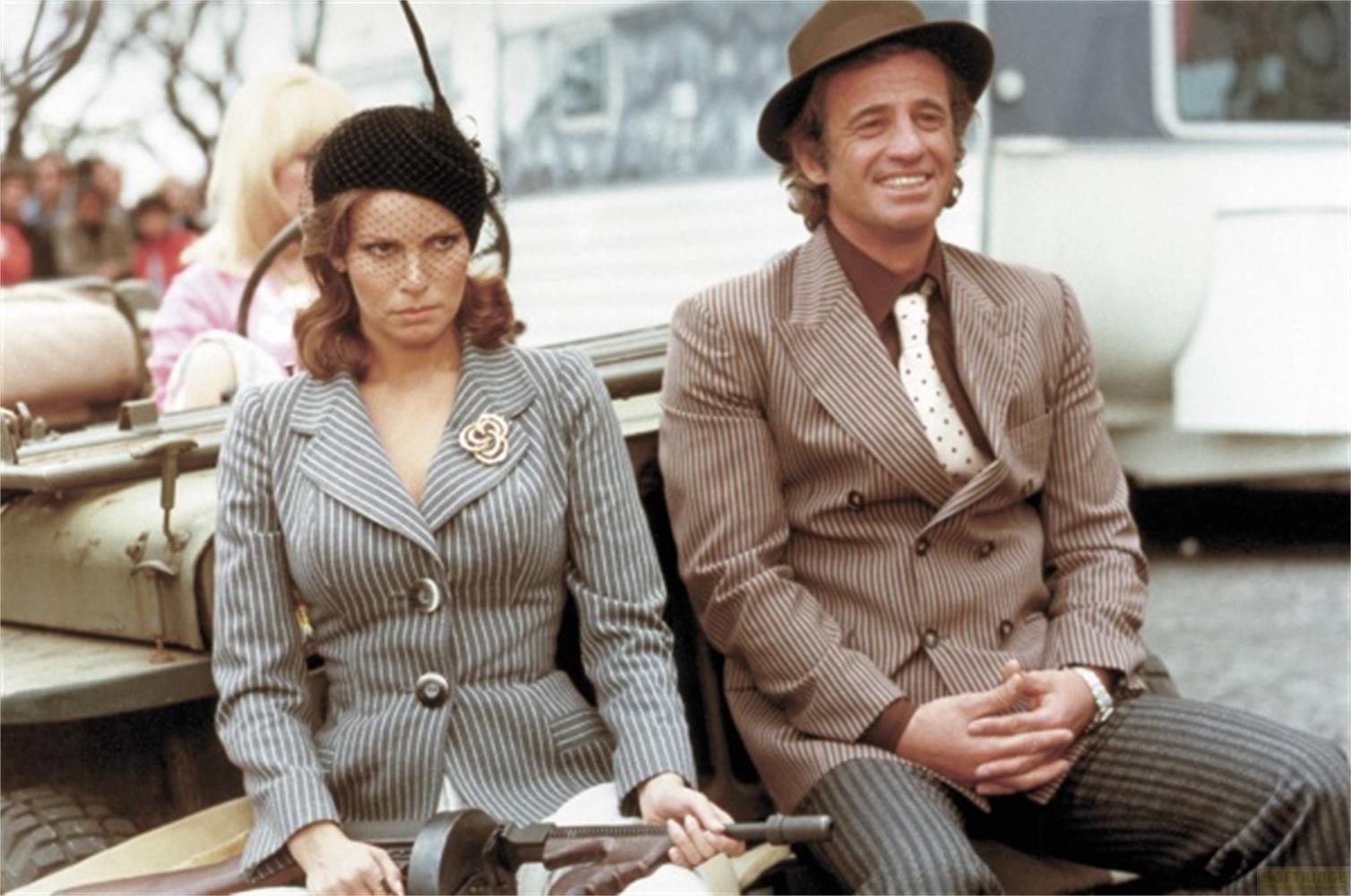 Donna con cappello e abito gessato accanto a uomo con cappello e abito.