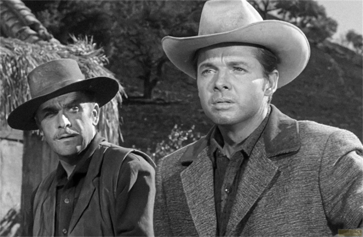 Due uomini con cappelli e abiti western davanti a una struttura rustica.
