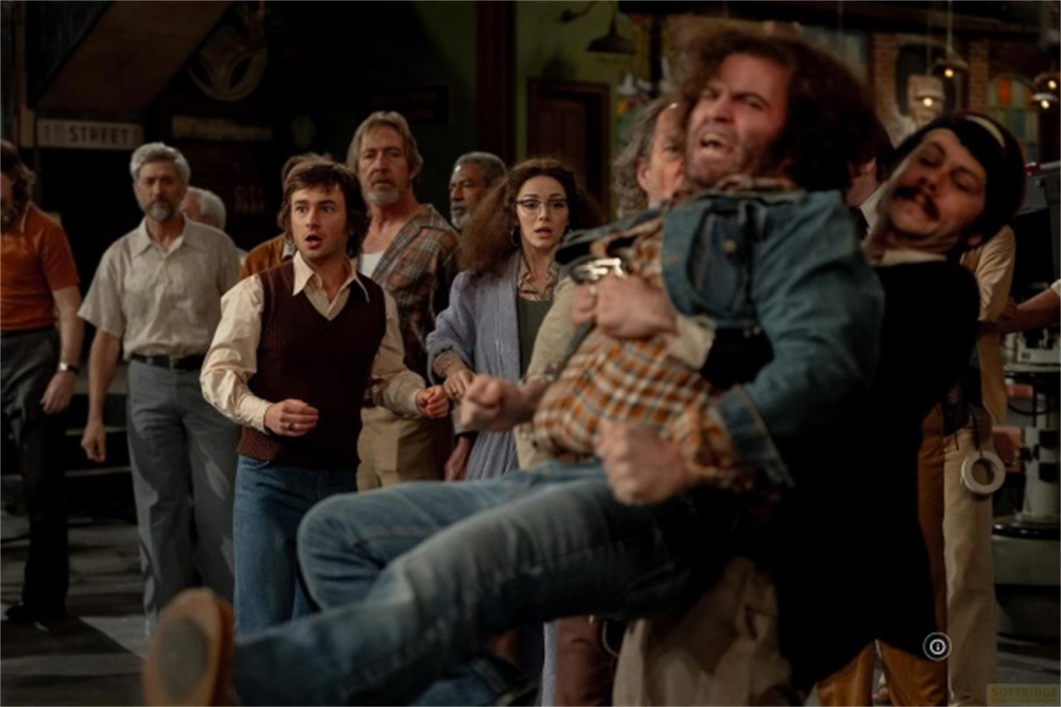 Un gruppo di persone vestite in abiti anni '70, alcuni sembrano eccitati, altri lottano.