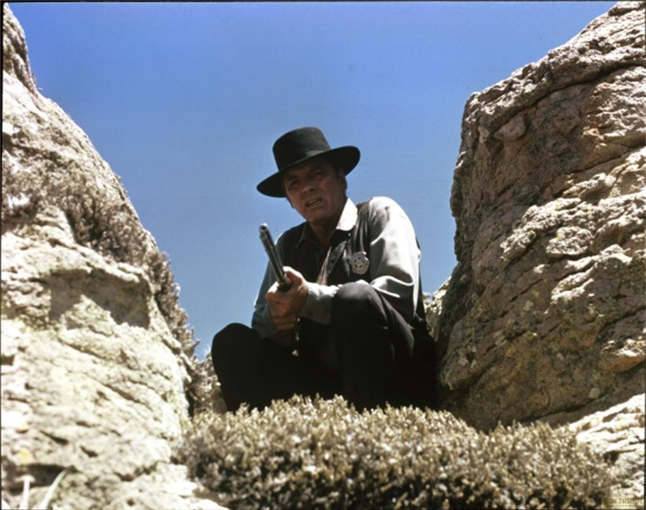 Un uomo con cappello e camicia neri tiene una pistola in un paesaggio roccioso.