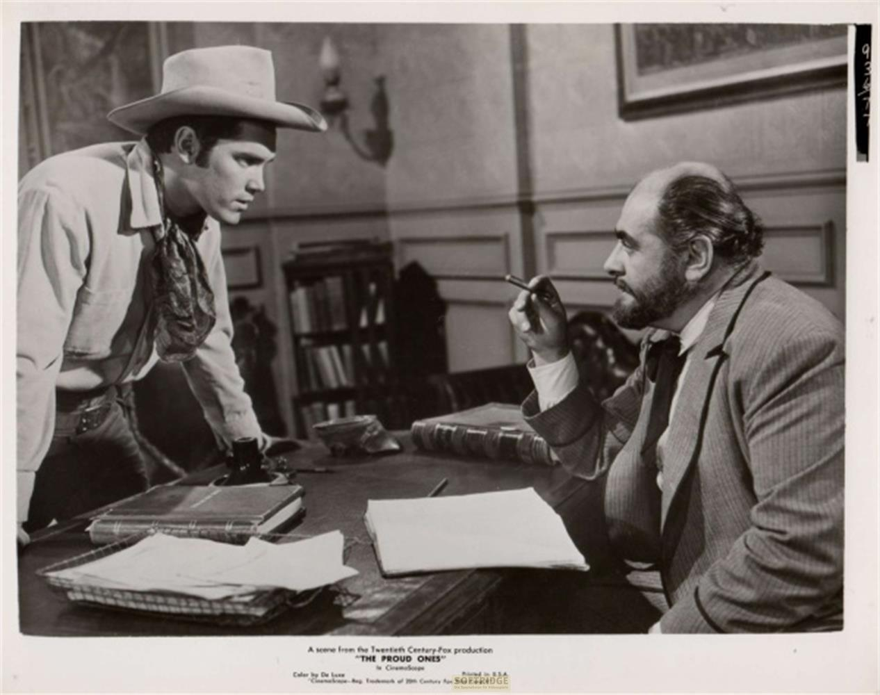 Un cowboy e un uomo con cappello siedono a una scrivania. Librerie sullo sfondo.