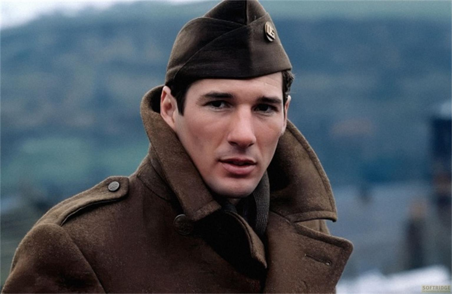 Ein Mann in Uniform mit braunem Mantel und Mütze steht vor unscharfem Hintergrund.