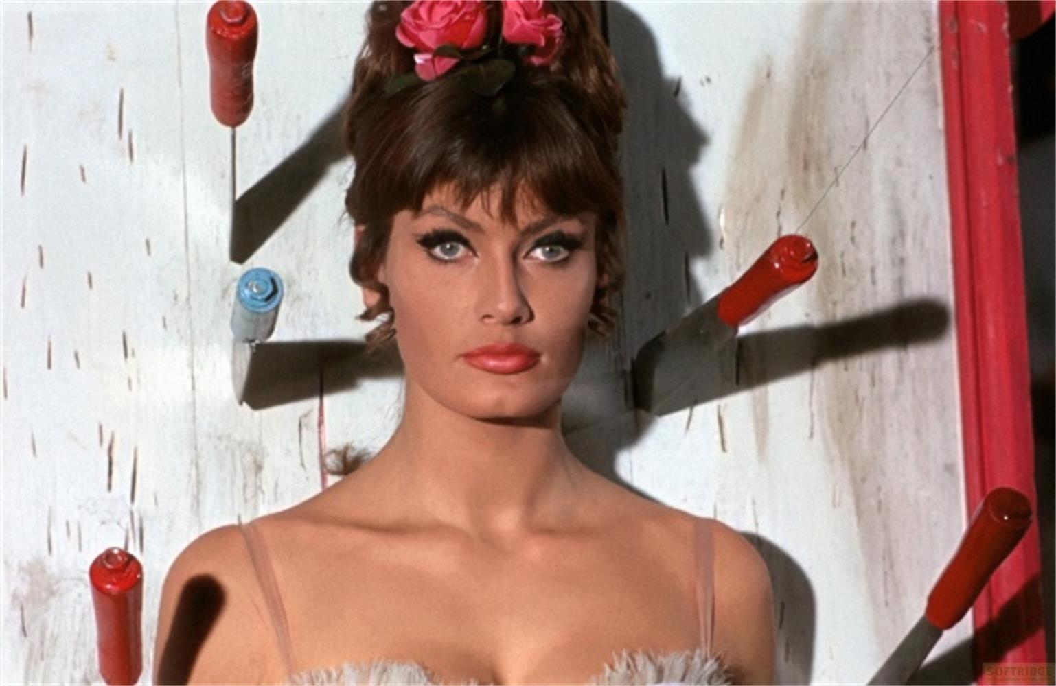 Una donna con fiori nei capelli sta davanti a un muro di legno con coltelli.