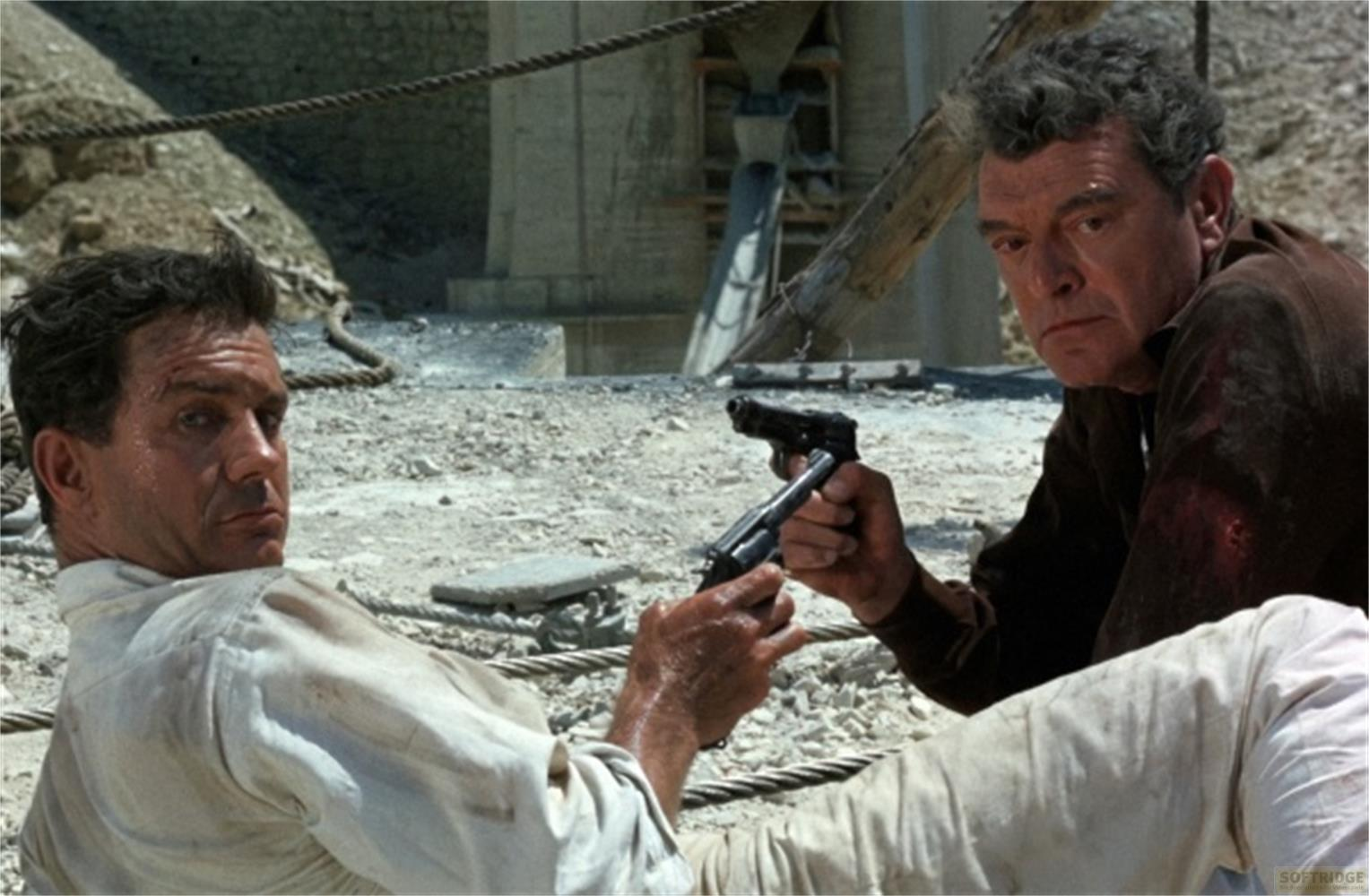 Due uomini all'aperto, uno tiene una pistola. Sembrano sporchi e tesi. Terreno roccioso.