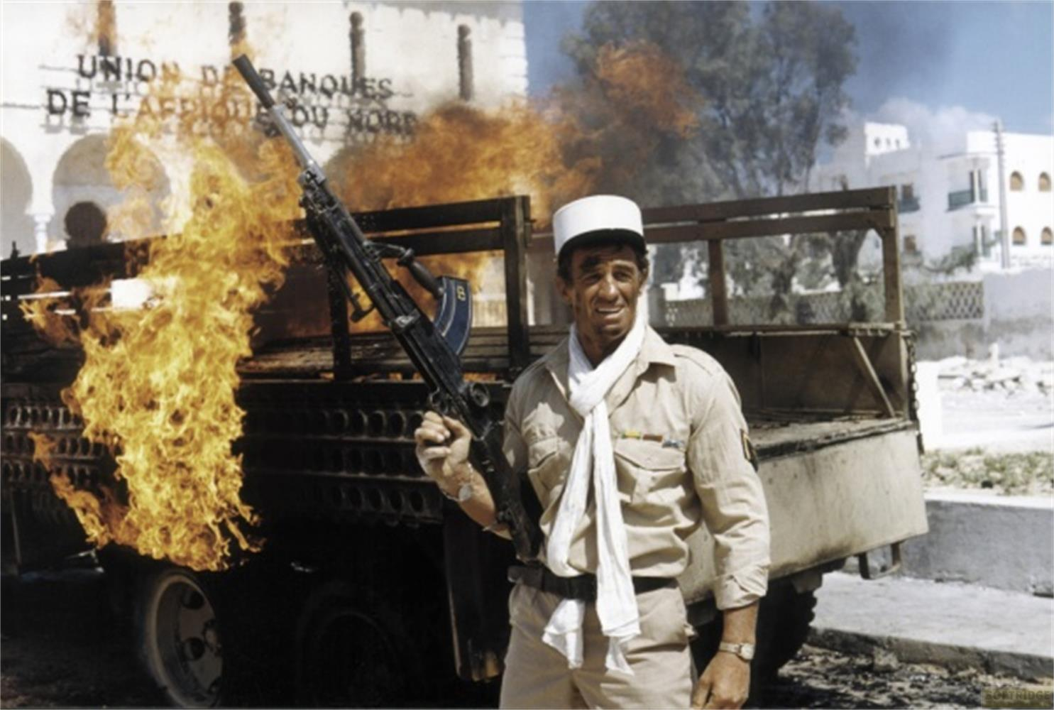 Uomo in uniforme tiene fucile accanto a veicolo in fiamme.