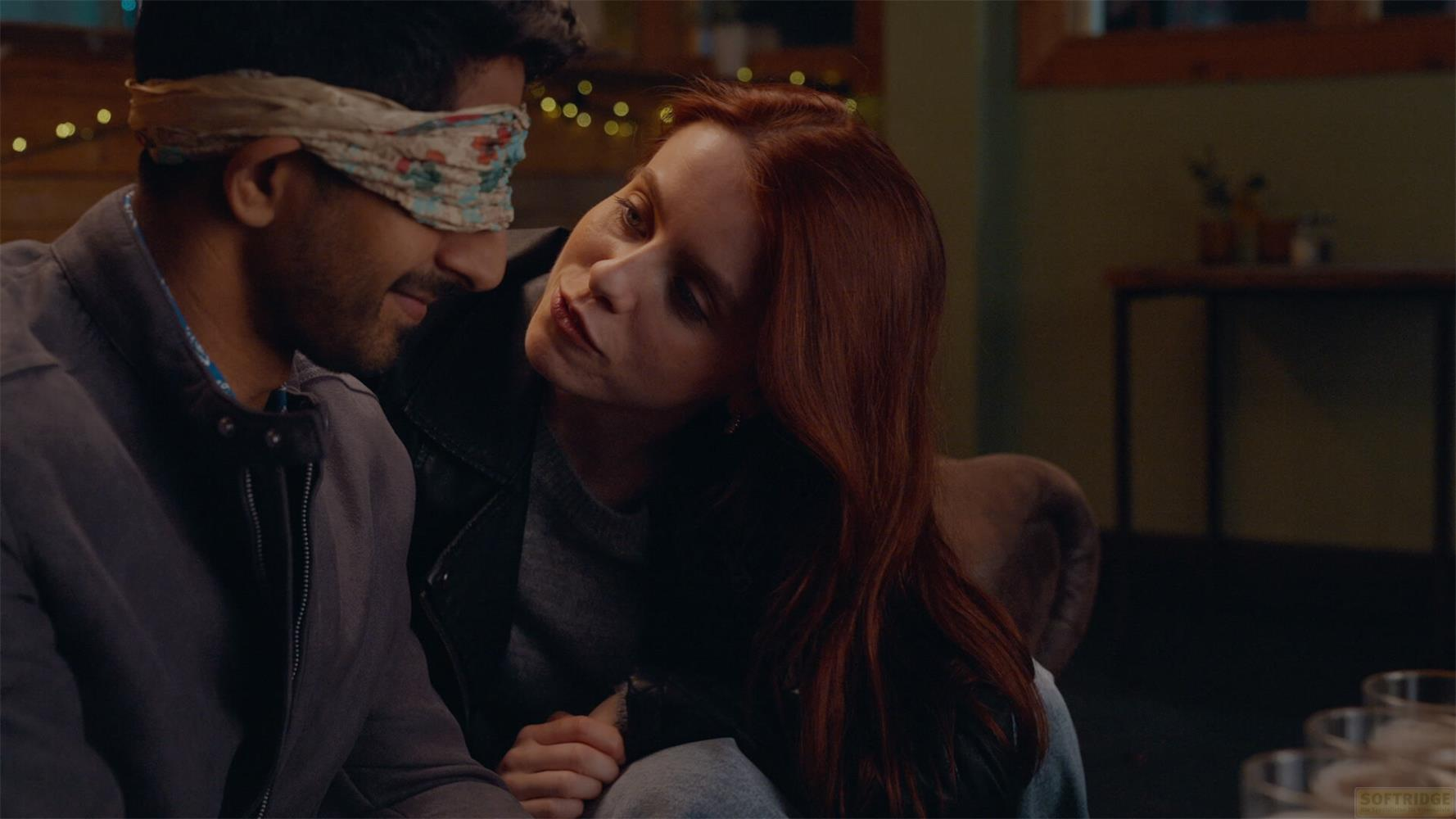 Homme bandeau sur les yeux regarde une femme rousse penchée vers lui.
