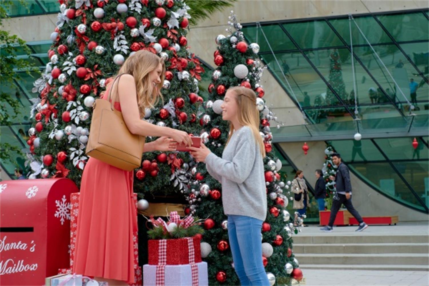 Due donne si scambiano regali davanti ad alberi di Natale decorati e una cassetta postale rossa.