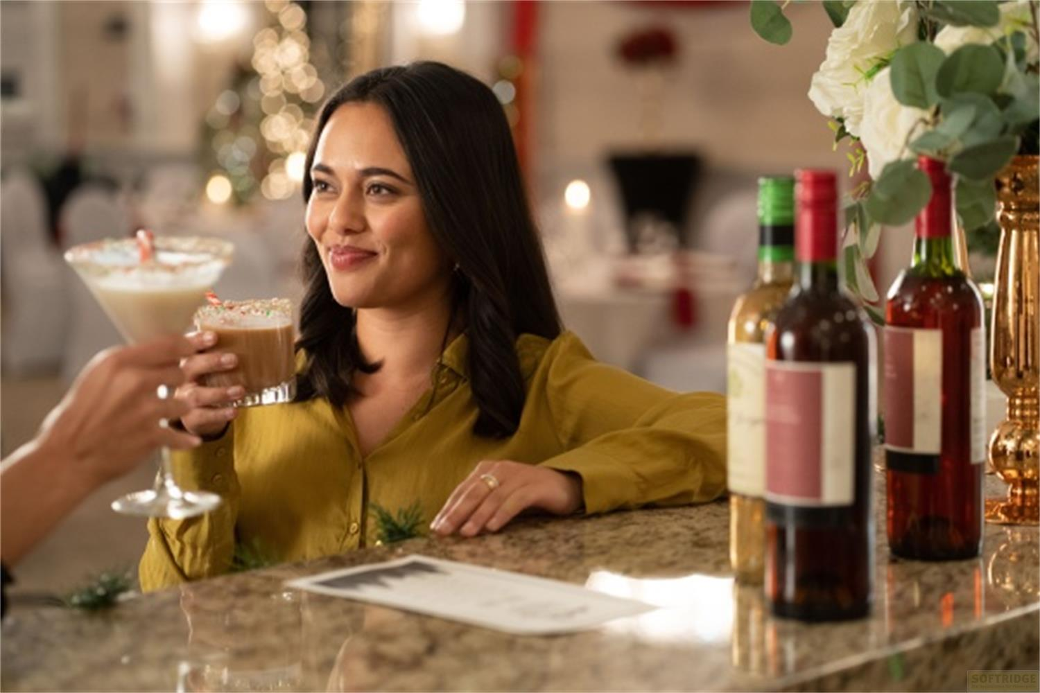 Una donna con una camicia gialla sorseggia un drink in un bar con decorazioni natalizie.