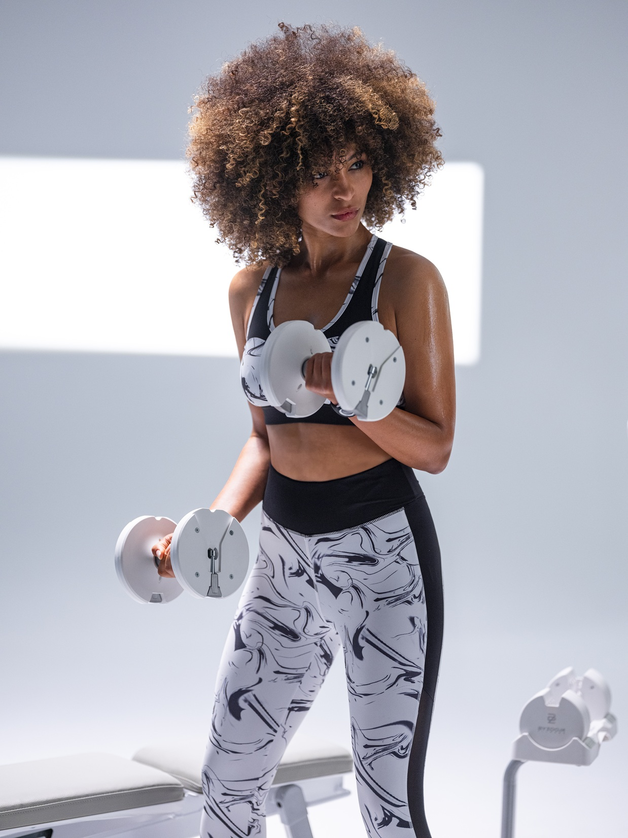 Eine Frau mit lockigem Haar hebt Hanteln. Sie trägt ein Sport-BH und gemusterte Leggings.