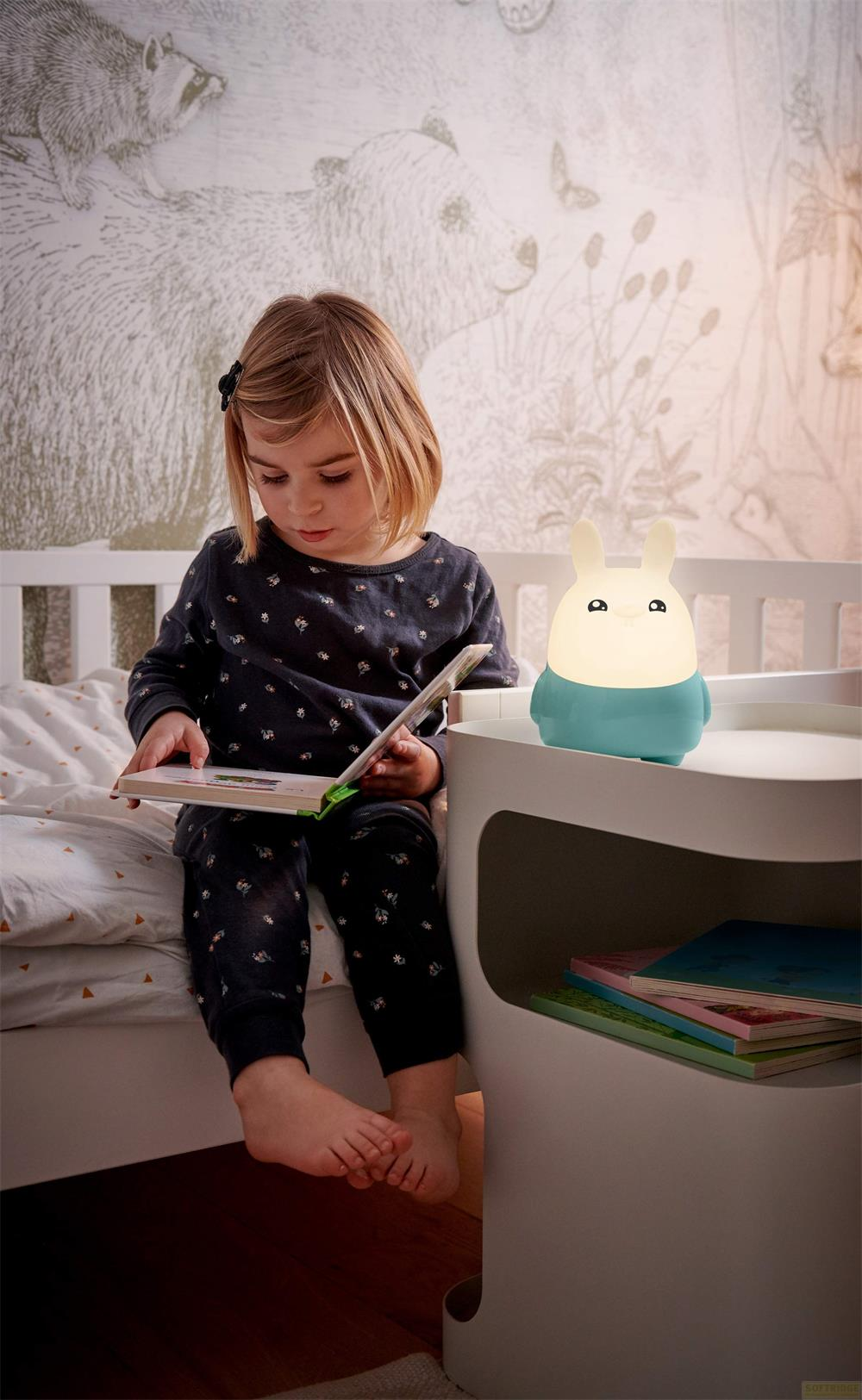 Una niña sentada en la cama lee un libro, con una lámpara de conejo brillante.