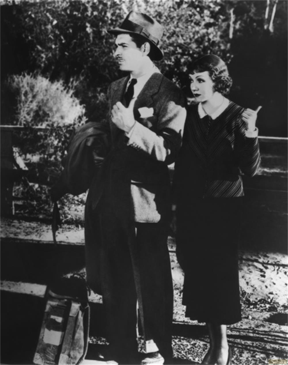 Homme en chapeau et costume à côté d'une femme en robe rayée. Une valise au sol.