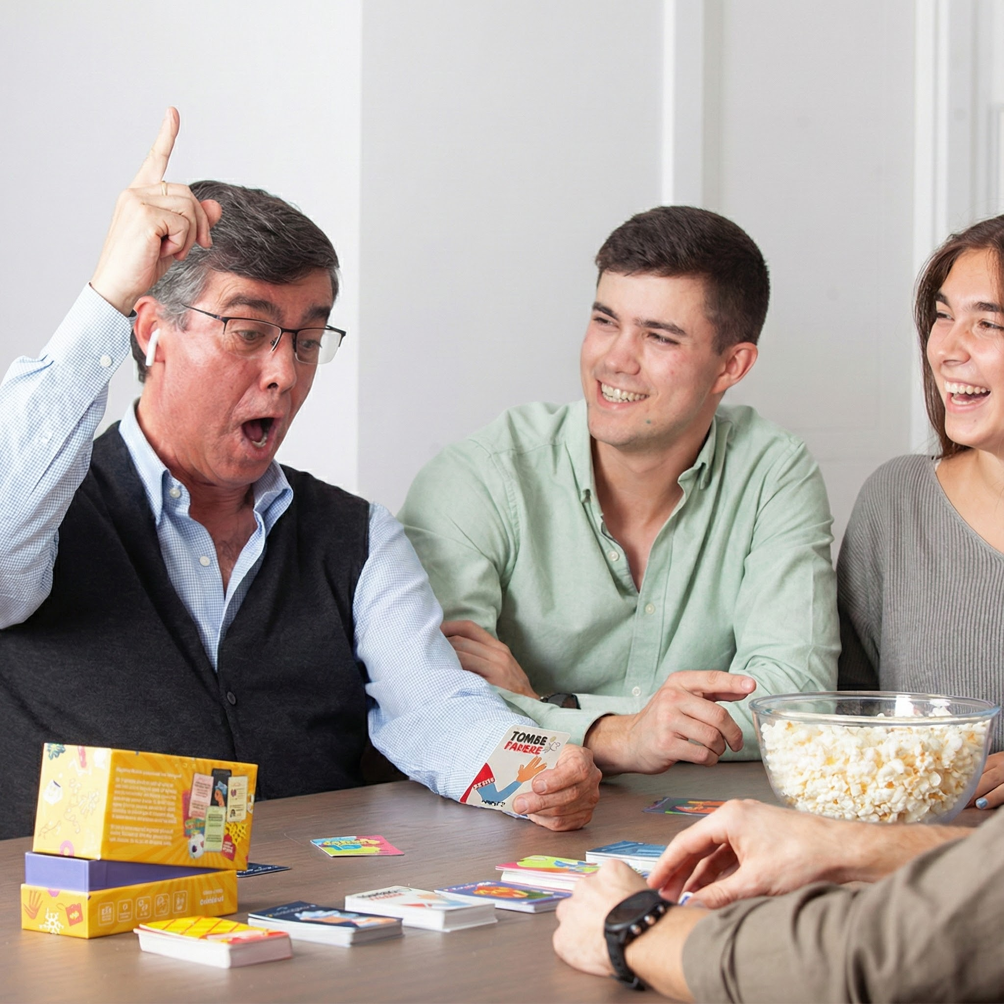Cuatro personas juegan a las cartas. Uno señala emocionado hacia arriba. Hay palomitas en la mesa.