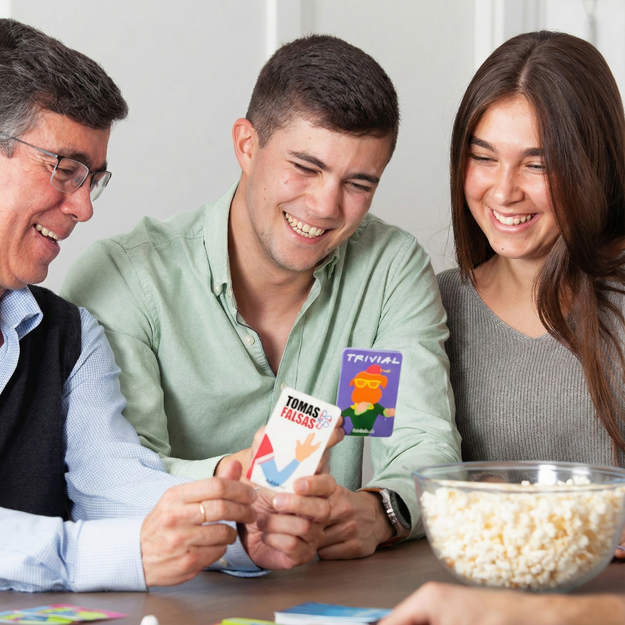 Tres personas riendo juegan a las cartas junto a un bol de palomitas.