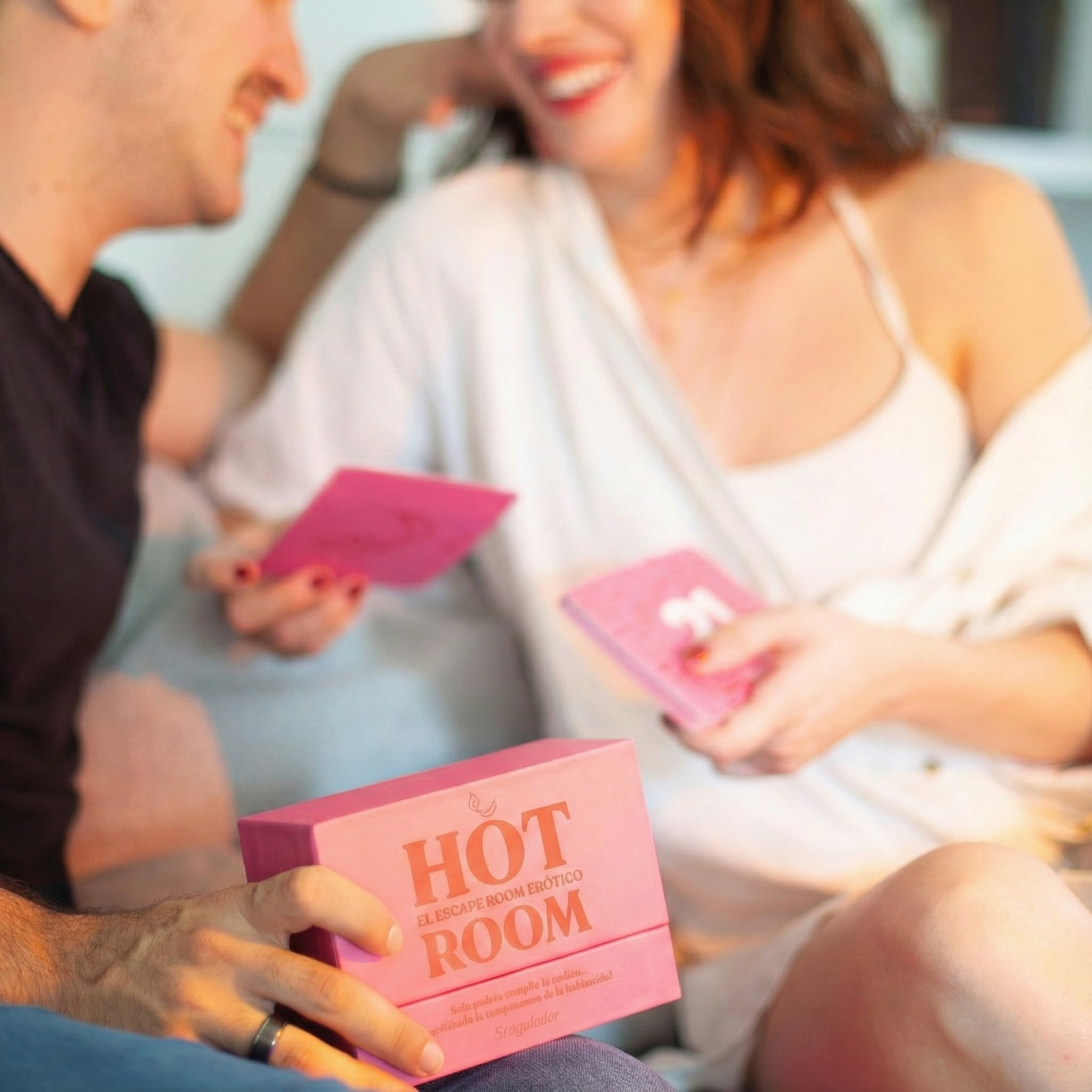 Una pareja ríe, sosteniendo cartas rosas. Una caja rosa con la etiqueta HOT ROOM está al frente.
