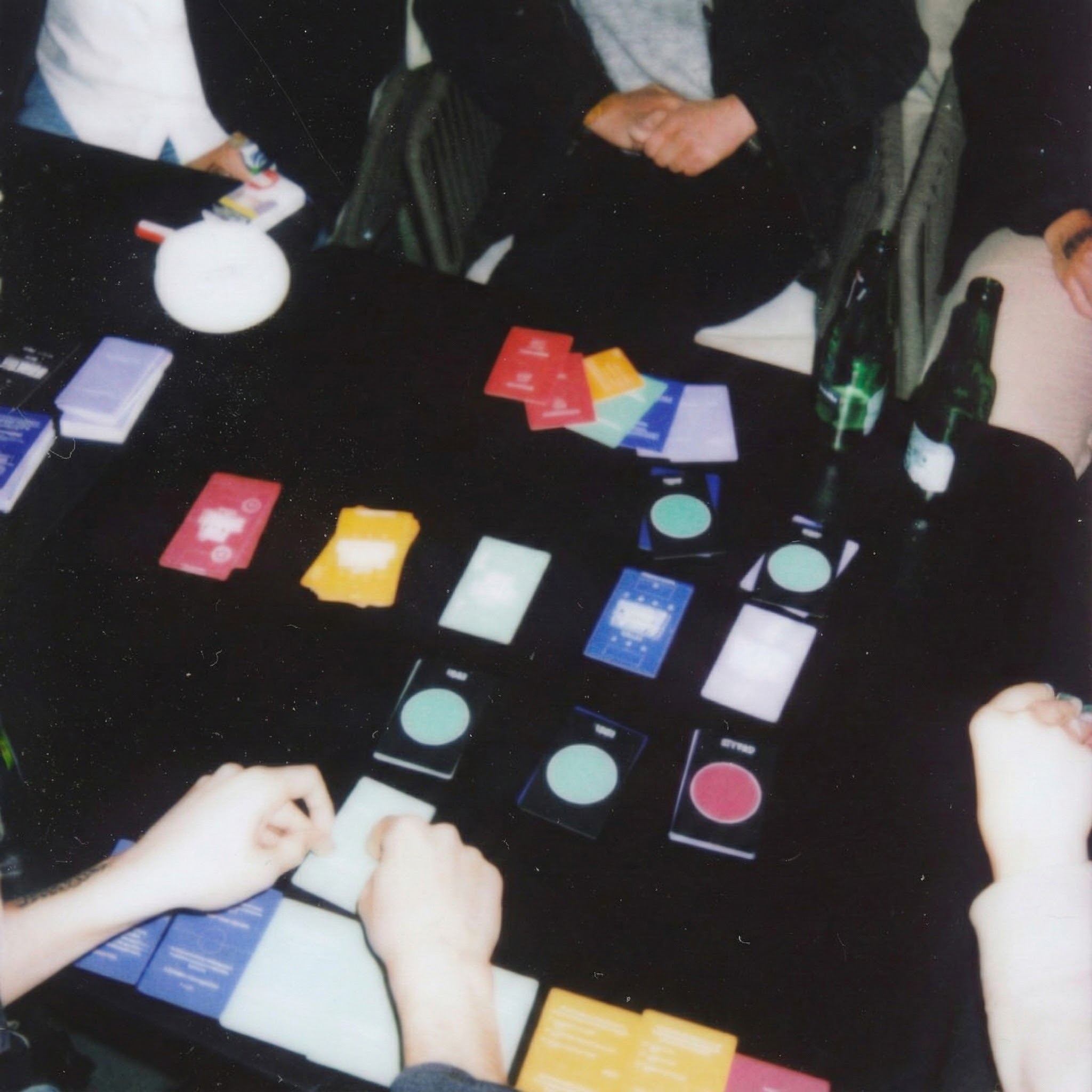 Personas juegan a las cartas sobre una mesa oscura con botellas y un objeto blanco.
